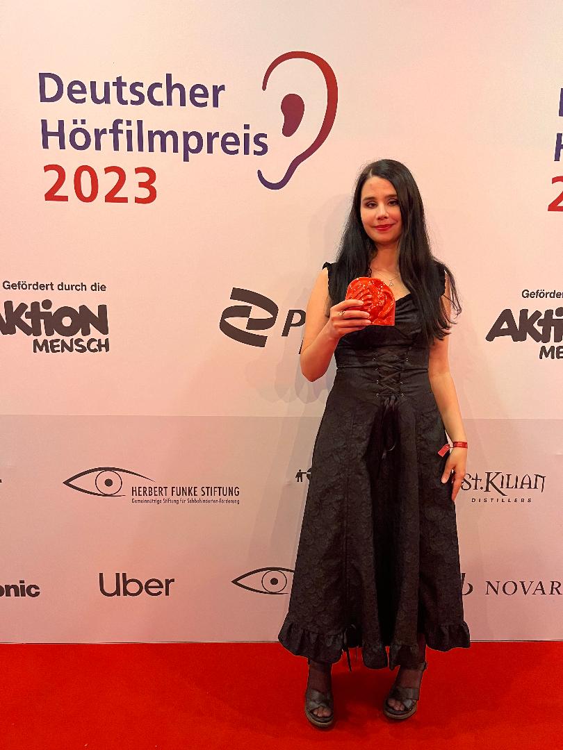 Johanna steht auf einem roten Teppich vor einer Wand, auf der groß "Deutscher Hörbuchpreis 2023" zu lesen ist. Sie hat offene, lange, schwarze Haare, traegt ein langes, schwarze Kleid im Gothic-Stil und haelt den roten Preis vor sich hoch.