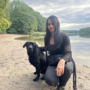 Johanna und ihr zukuenftiger Blindenfuehrhund Aris, ein schwarzer Labrador, sitzen nebeneinander an einem See. Beide schauen in die Kamera. Aris ist relativ klein und hat ein baeriges Gesicht. Johanna laechelt gluecklich. Ihre langen schwarzen Haare fallen vorne ueber ihre Schulter.